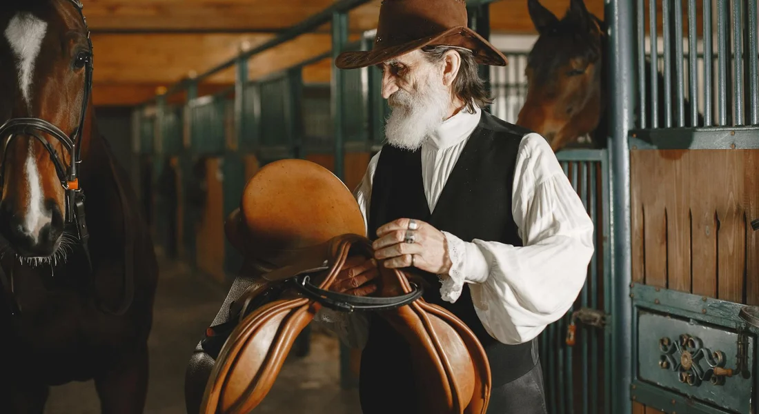 Older man with a beard wearing a cowboy hat and vest, handling a saddle inside a horse stable with horses in stalls.