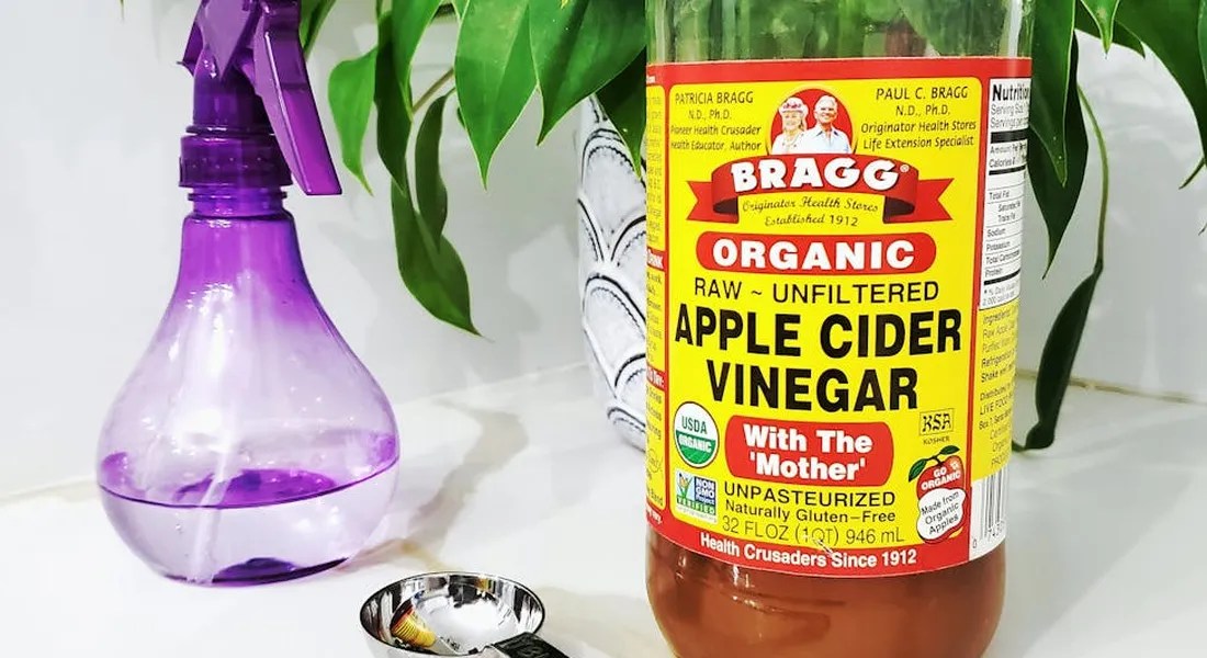Bragg Organic Apple Cider Vinegar bottle labeled 'With The Mother' beside a purple spray bottle on a white surface