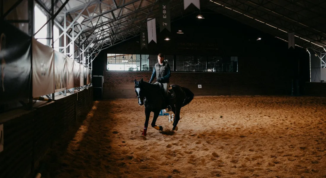 Inside a dim indoor riding arena, a rider on a horse walks slowly along the sandy floor.