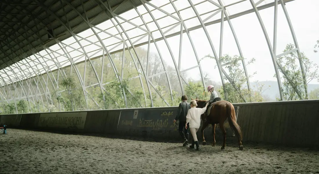 Two people assist a horse inside a large indoor riding arena, demonstrating calm, cooperative training in a controlled environment.