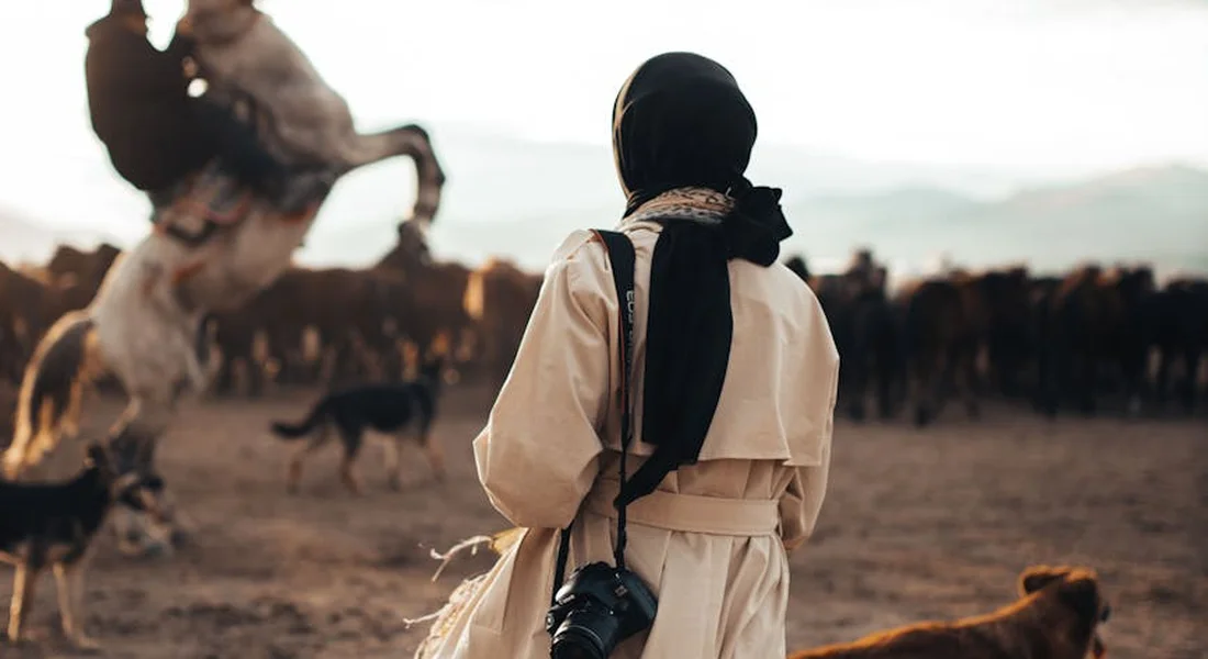 Back view of a person wearing a scarf and beige coat with a camera around their neck, observing a herd of horses in a dusty, open landscape.