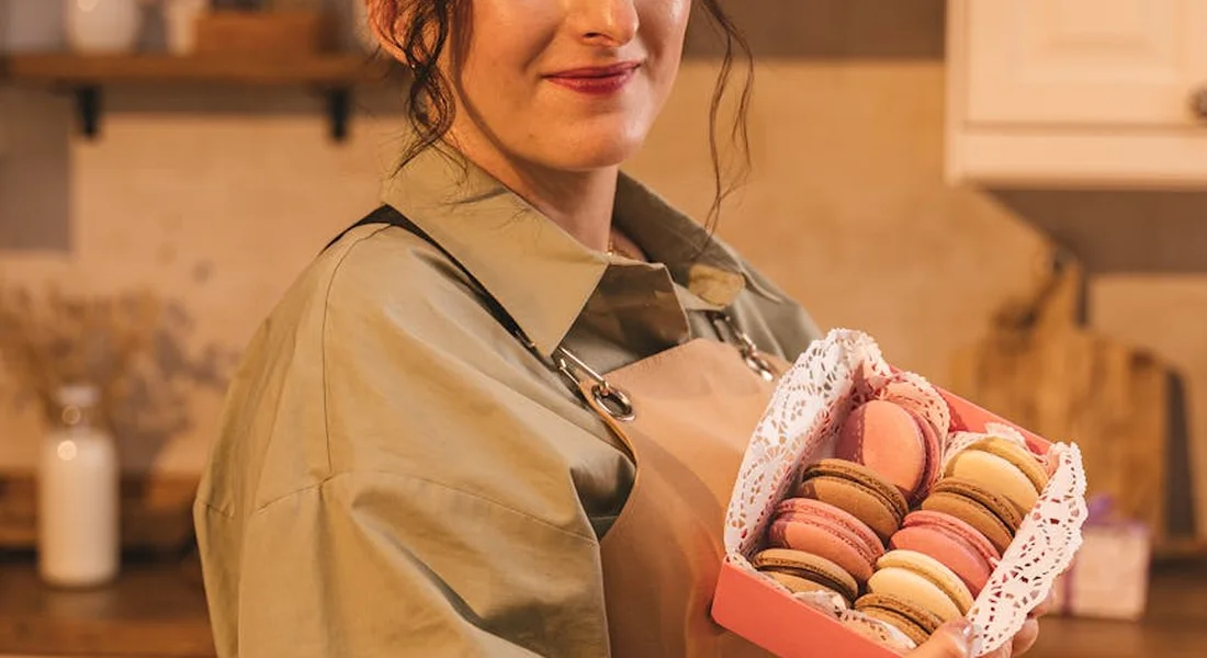 A person in an apron holds a box of colorful macarons in a cozy kitchen.