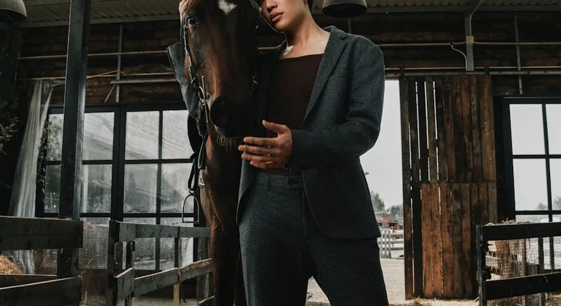 Person in a dark blazer stands beside a horse inside a stable, gently touching the horse.