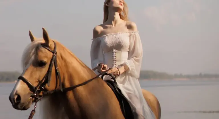 A woman wearing an off-the-shoulder white dress rides a light-colored horse along a beach, with water and a pale sky in the background.