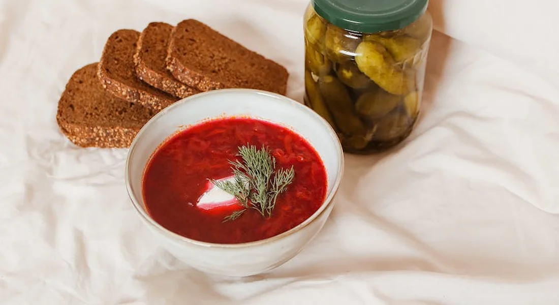 A bowl of red beet soup garnished with dill, accompanied by dark rye bread slices and a jar of pickles on a light cloth.