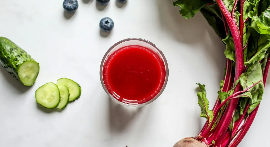 Top-down flat lay of a glass of red beet juice surrounded by cucumber slices, blueberries, and fresh beet greens with red stems, illustrating beets as a treat alongside other snack options.