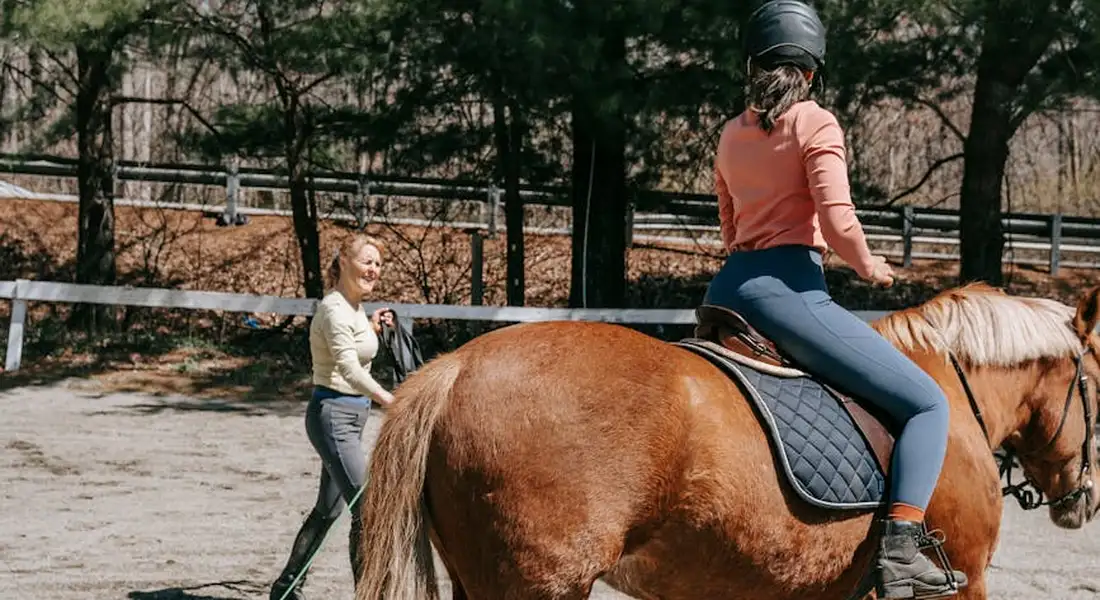 Rider on a horse practicing in an outdoor arena while a trainer watches from the ground