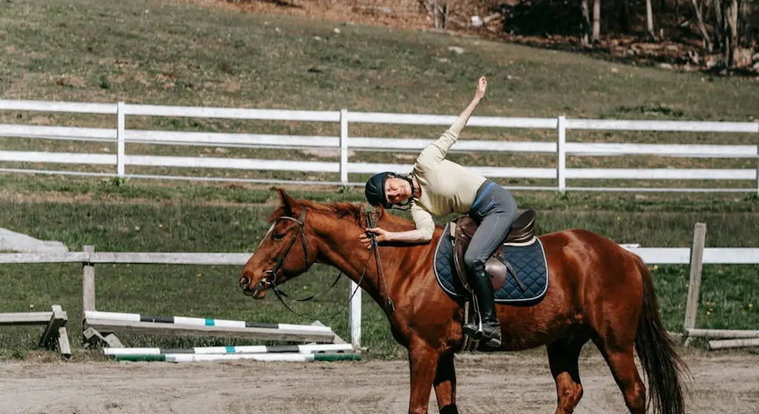 Rider on a brown horse in an outdoor riding arena, performing a mounted stretch with the rider leaning sideways and one arm raised.