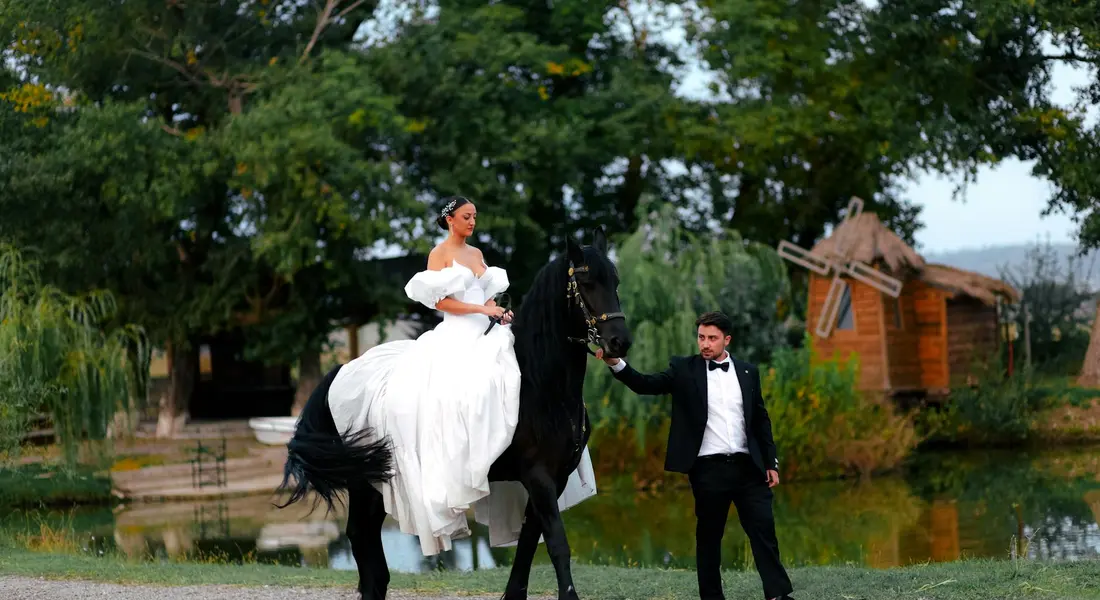 A bride in a white dress rides a black horse, guided by a man in a tuxedo in a park setting near a pond.