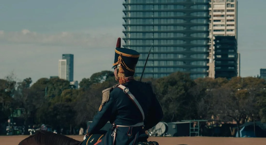 Back view of a rider in ceremonial uniform on a horse, with city buildings in the background.