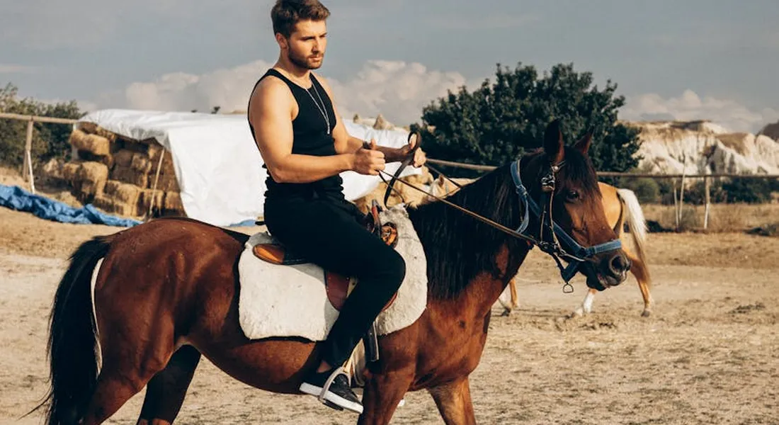 Rider on a brown horse in an outdoor arena; man wearing a black sleeveless shirt and dark jeans; white saddle pad; fenced corral with hay bales and trees in the background.
