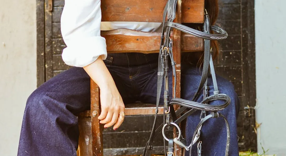 A person in white shirt sleeves and dark pants sits on a wooden chair with horse tack (bridle and reins) draped over their lap, symbolizing preparation and collaboration in equine care.