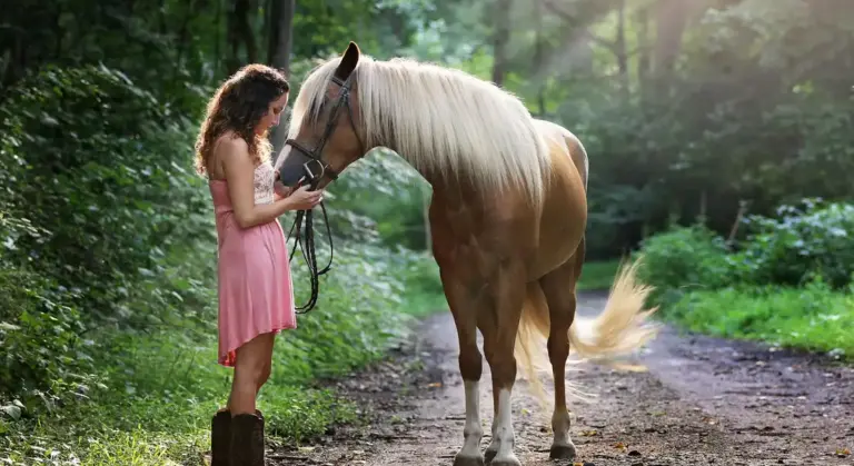 A woman in a pink dress standing on a forest path, gently holding the reins of a large light-colored horse with a white mane.