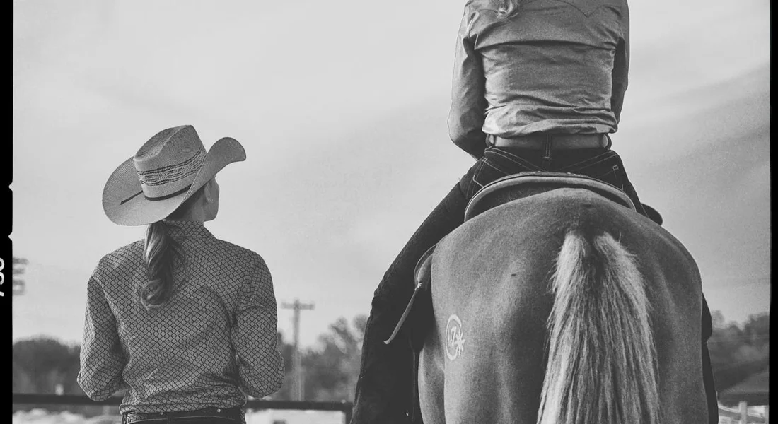 A rider on a horse seen from behind with a second person wearing a wide-brim hat standing nearby; black-and-white photograph.