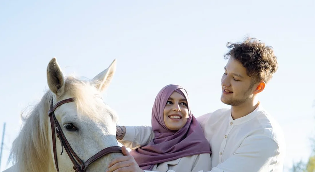 A smiling couple stands beside a calm white horse outdoors; the woman wears a pink headscarf, illustrating gentle horse-human bonding.