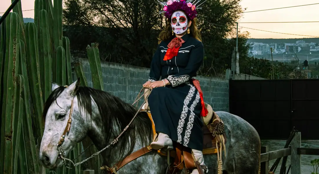A rider wearing Day of the Dead makeup and a decorative outfit sits atop a gray American Paint Horse in an outdoor corral with tall cacti and a low stone wall in the background.