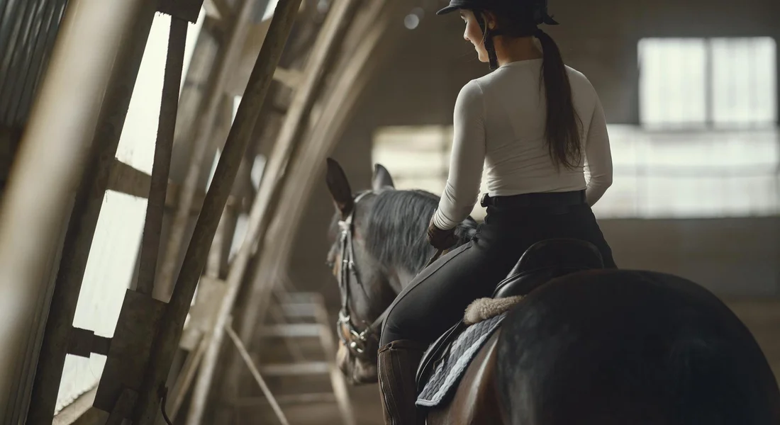 Rider in a white long-sleeve shirt wearing a helmet sits on a dark horse inside an indoor riding arena, with wooden beams along the side.