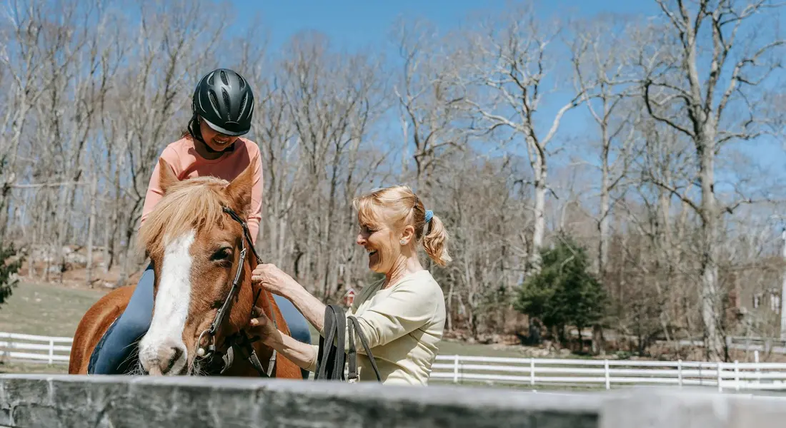 Child rider on a horse with an adult trainer beside them in an outdoor arena, blue sky, leafless trees, and a white fence in the background.