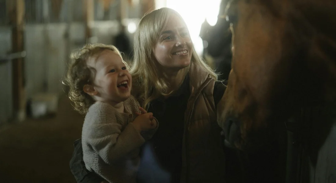 A smiling woman holding a child beside a horse in a dimly lit stable.