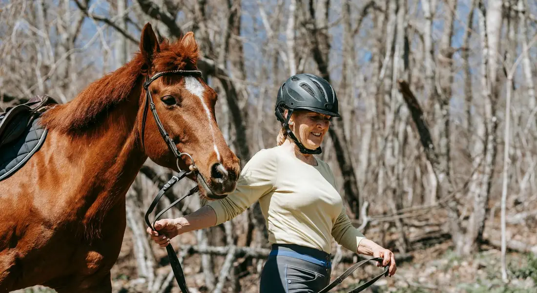 Rider wearing a helmet and light shirt guiding a calm horse along a forest trail.