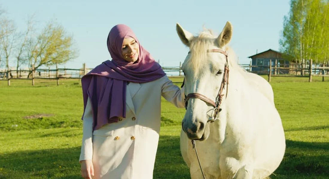 A woman wearing a purple hijab stands beside a white horse in a sunlit grassy field.