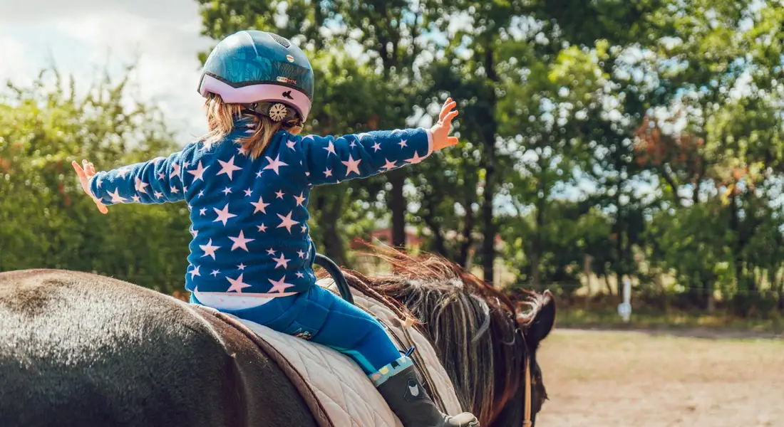 A child wearing a star-pattern blue jacket and helmet sits on a dark horse with arms extended, practicing balance during outdoor groundwork.
