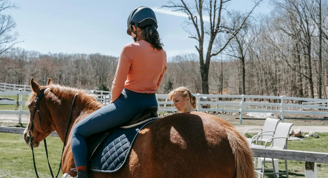 A rider wearing a helmet and orange shirt sits atop a brown horse in an outdoor arena, while a trainer stands nearby to guide groundwork.