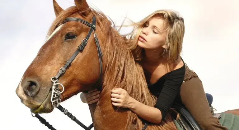 A rider sits on a brown horse with a bridle, leaning close to the horse’s neck as she holds the reins.
