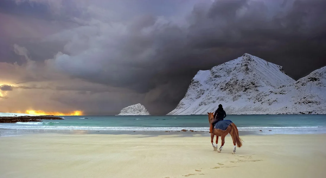 A rider on horseback walking along a sandy beach with dark, stormy clouds and snow-covered mountains in the distance.
