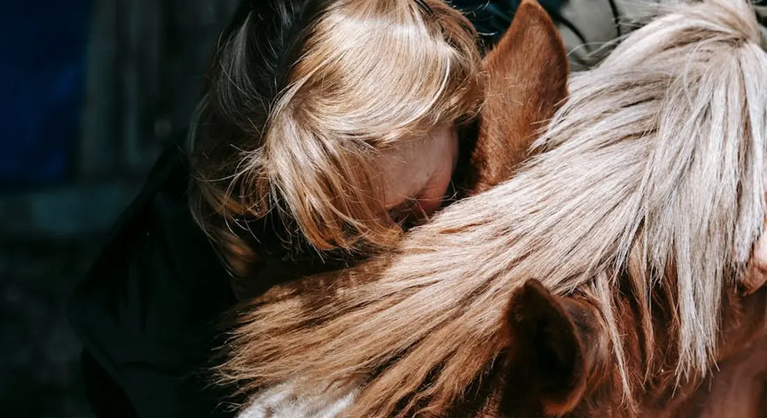 Close-up of a blond-haired person embracing the mane of a light brown horse