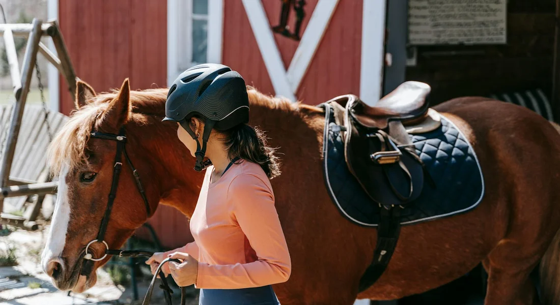 Person wearing a helmet standing beside a saddled horse near a red barn, preparing for riding.