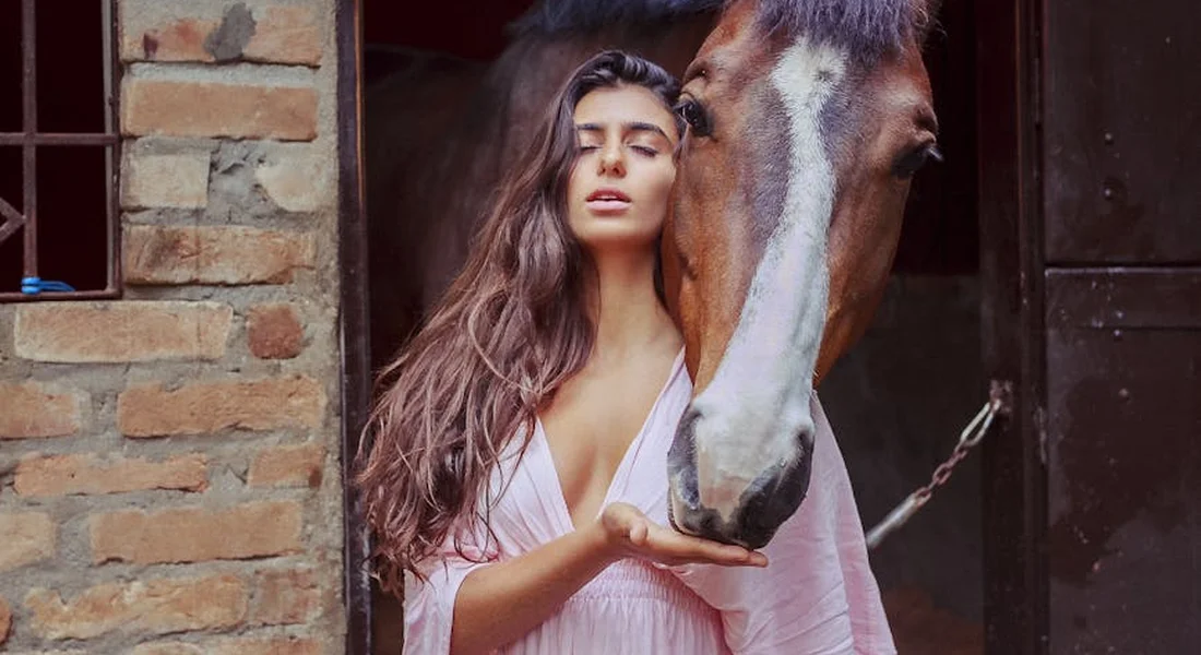 A woman with long dark hair in a light pink robe stands at a stable doorway, touching noses with a horse.