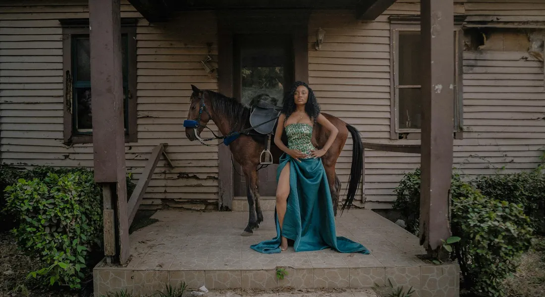 A woman in a turquoise dress stands beside a horse on a weathered porch, with the horse's side visible for flank health observation.