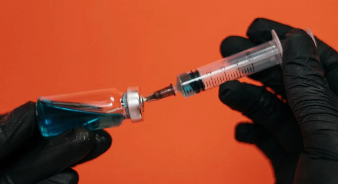 Close-up of gloved hands preparing a syringe and vial with blue liquid against an orange background, illustrating the injection technique for horses.