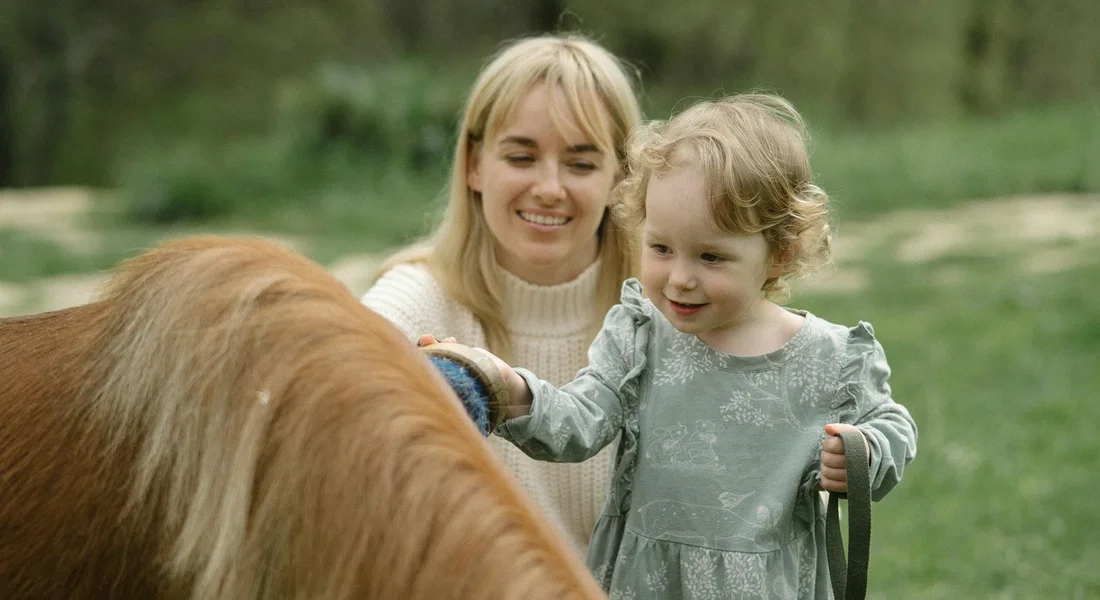 A woman and a young girl groom a horse's mane outdoors, smiling as they work with a brush.