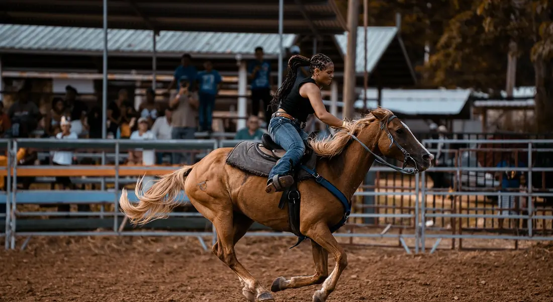 A rider on a brown horse galloping across a dusty arena, with fencing and spectators in the background.