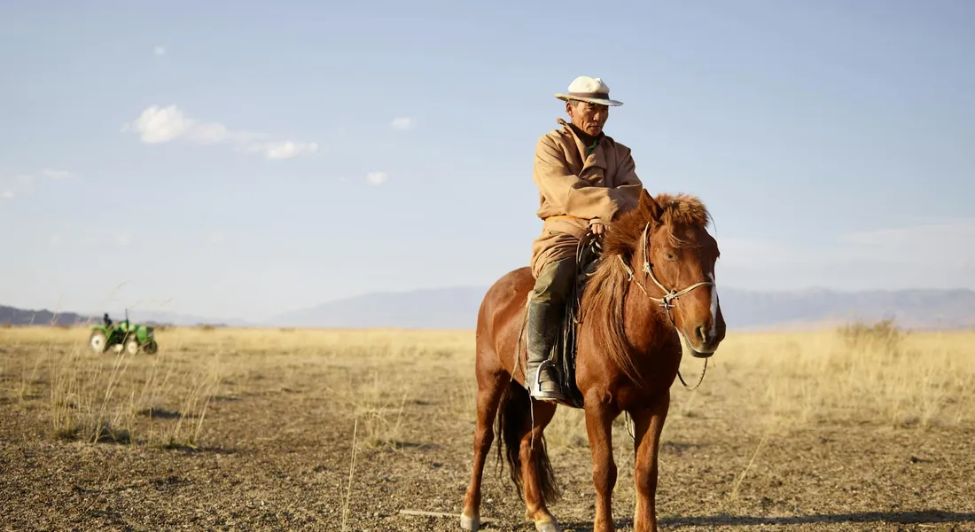 A man wearing a wide-brimmed hat rides a brown horse across a dry, open field with distant hills.