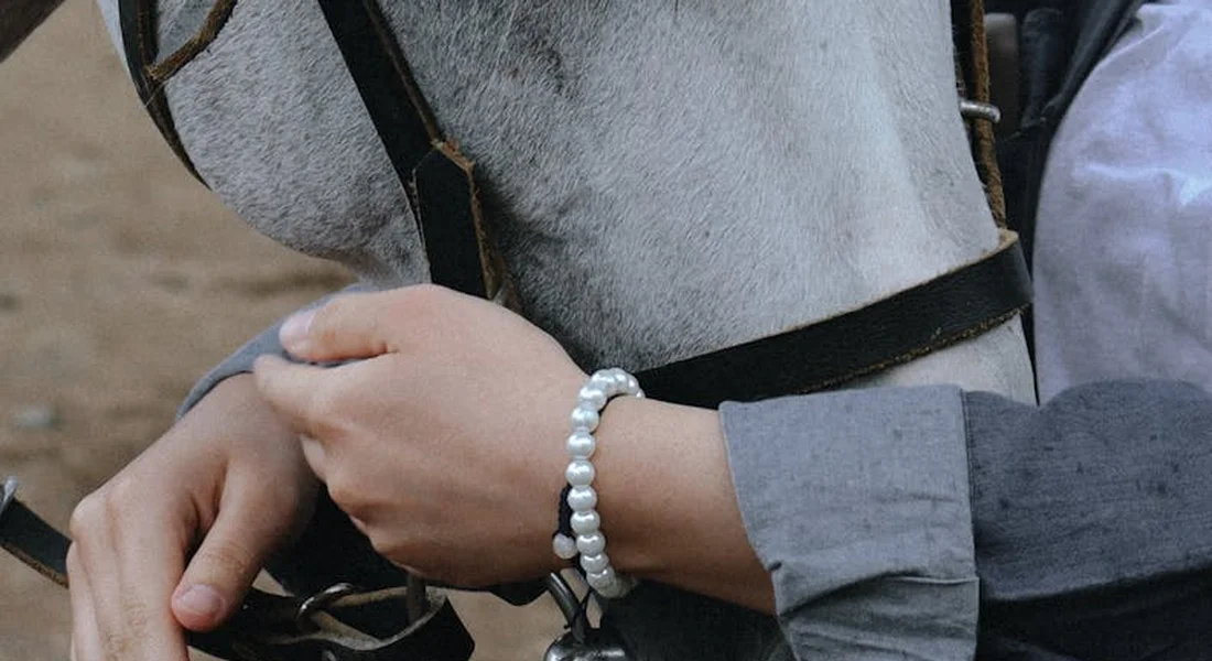 Close-up of a rider's hands gripping horse reins, wearing a pearl bracelet, with part of the horse and saddle visible.