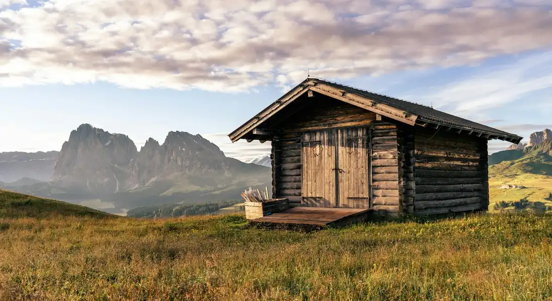 A rustic wooden horse run-in shed on a grassy hillside with mountains in the distance.