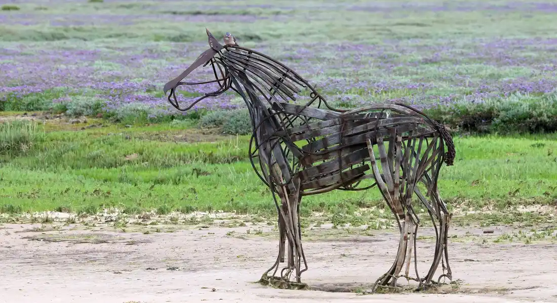 Metal skeletal horse sculpture standing on a dirt path with green grass and purple flowers in the background.