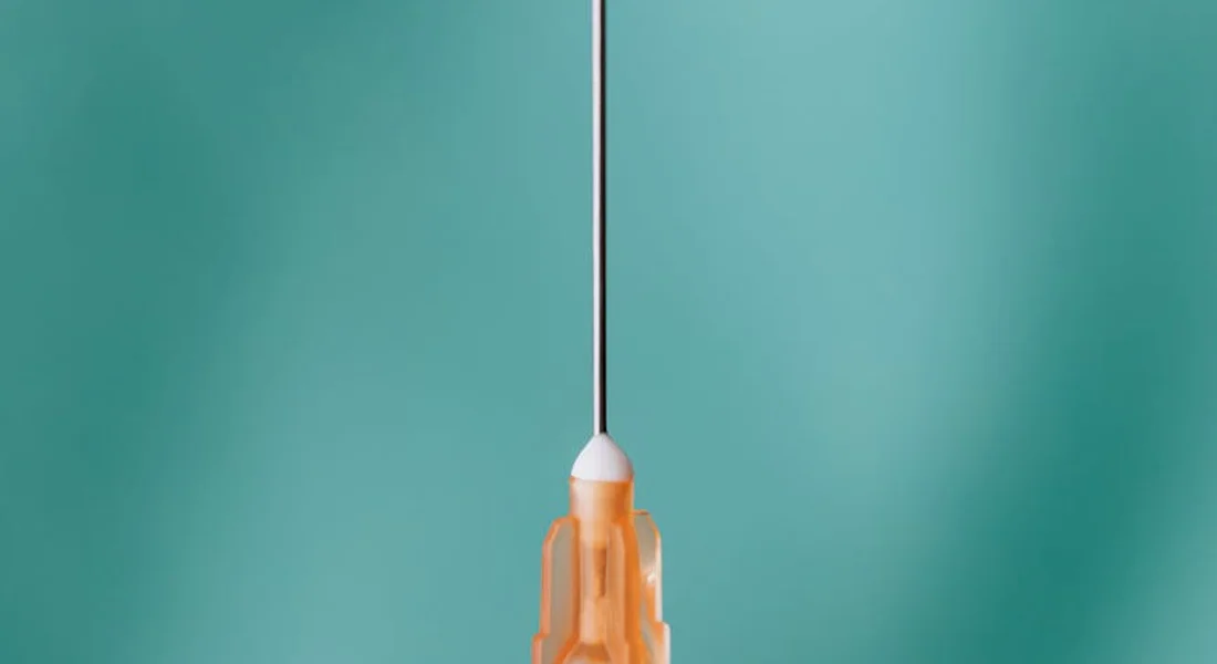 Close-up of a syringe needle with an orange plunger against a teal background.