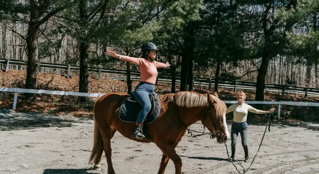 Rider wearing a helmet and pink top sits tall on a brown horse in an outdoor arena, while a trainer on the ground holds a longe line nearby