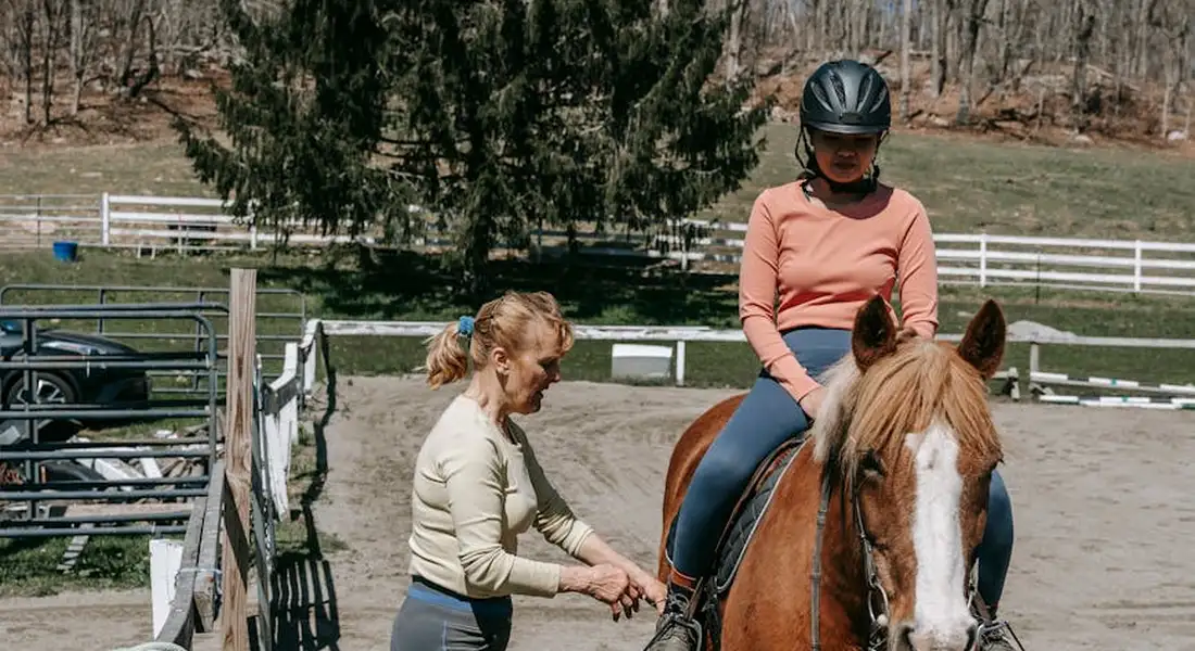 A rider wearing a helmet sits on a horse in an outdoor arena while a trainer on foot guides a stretching exercise at the horse's shoulder, illustrating flexibility and mobility work.