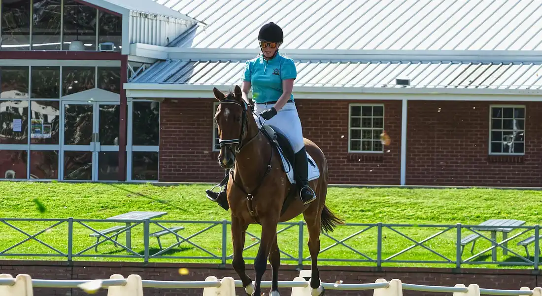 Rider on a brown horse wearing a helmet and teal shirt, moving through an outdoor riding arena with a brick building in the background.