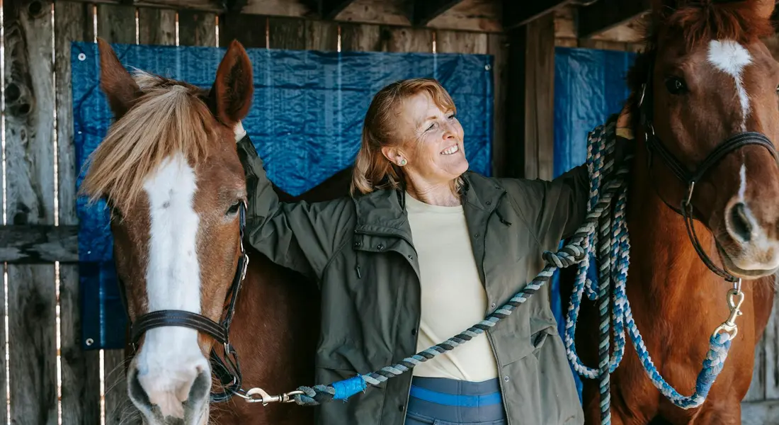 A woman in a jacket standing between two horses in a stable, smiling and holding their lead ropes.