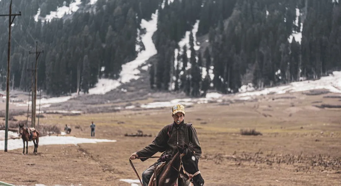 A rider on horseback in a wide, open winter pasture with patches of ice and snow, and snow-dusted mountains in the background; distant figures on horseback are visible.