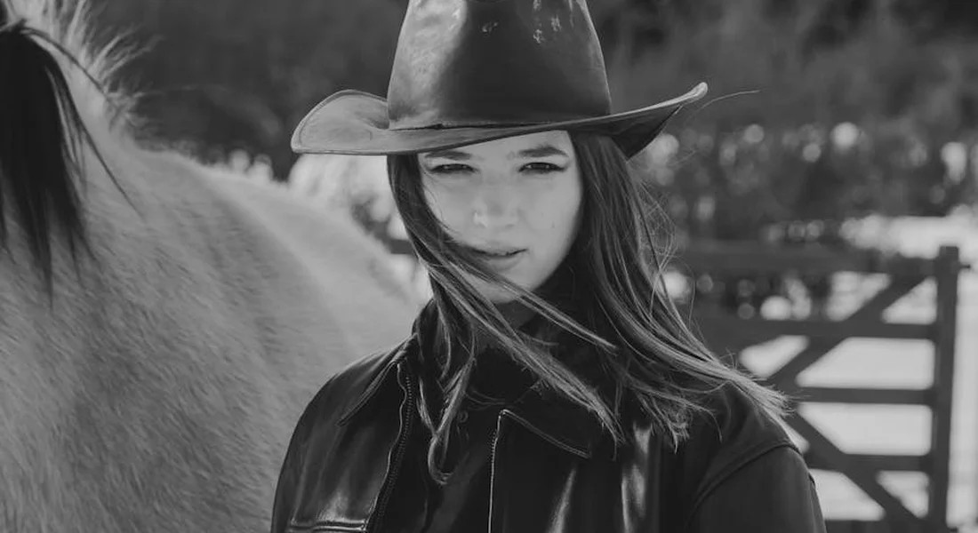 Black-and-white photo of a woman wearing a wide-brimmed hat standing beside a horse in an outdoor setting.