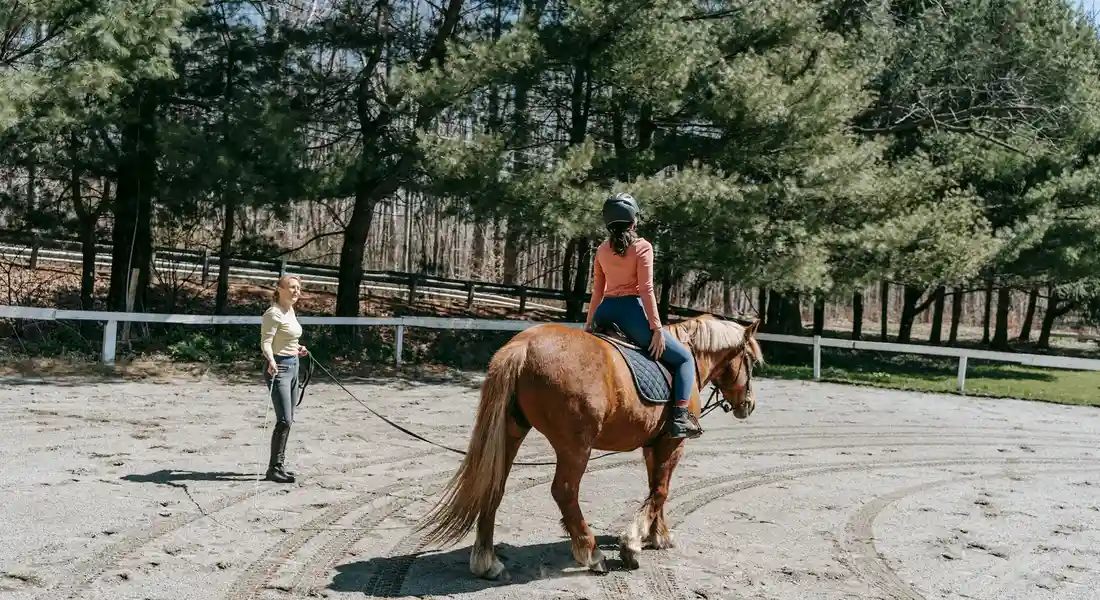 A rider on a chestnut horse in an outdoor riding arena, with a trainer standing nearby holding the horse's lead.
