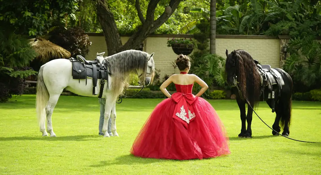 Two horses stand on a sunny, well-kept lawn flanking a person in a bright red ball gown, with trees and a garden setting in the background.