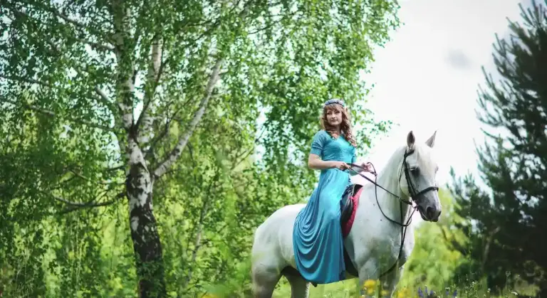 A rider in a blue dress sits astride a white horse in a sunlit forest, demonstrating calm, controlled handling.